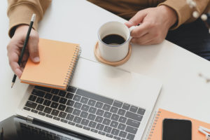 Person holding coffee cup and notebook in front of laptop